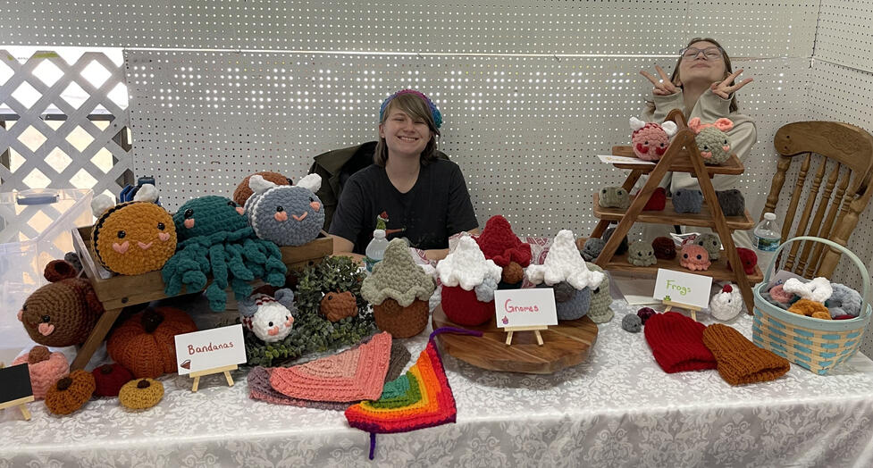 Two people smiling at a table covered in animal inspired plushies including bumble bees, octopi, gnomes, and frogs.