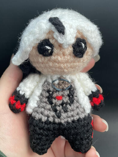 A crochet doll with white hair that has a dark grey streak in the bangs in a white coat with black and red cuffs and a grey shirt.
