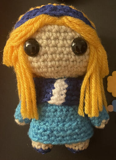 A crochet doll with yellow hair and a dark blue headband in a light blue dress with dark blue accents.