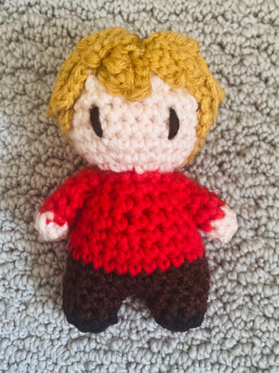 A crochet doll with dark yellow hair, a red shirt, and brown pants.