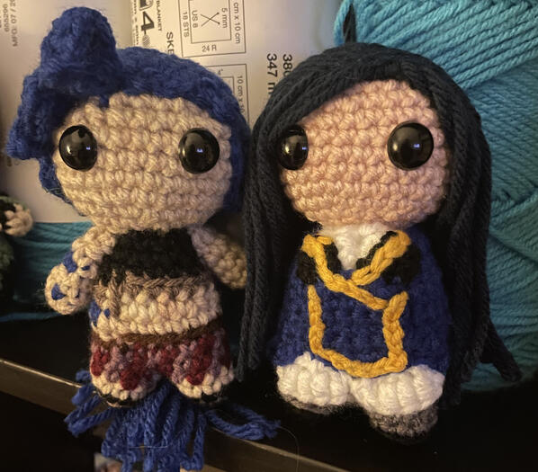 Two small crochet dolls stand next to each other on a bookcase. both have blue hair, but the character on the left has a lighter shade that is in braids. The doll on the right has straight hair and is in a military uniform.