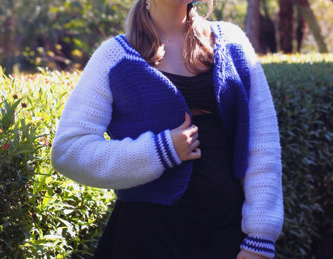 A girl standing in front of bushes wearing a black dress, glasses, and a hat modelling a hand-crocheted letterman jacket in white and mid-tone blue.