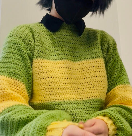 A person in a black wig wearing a black mask in a green, hand crocheted sweater with a large yellow stripe across the center.